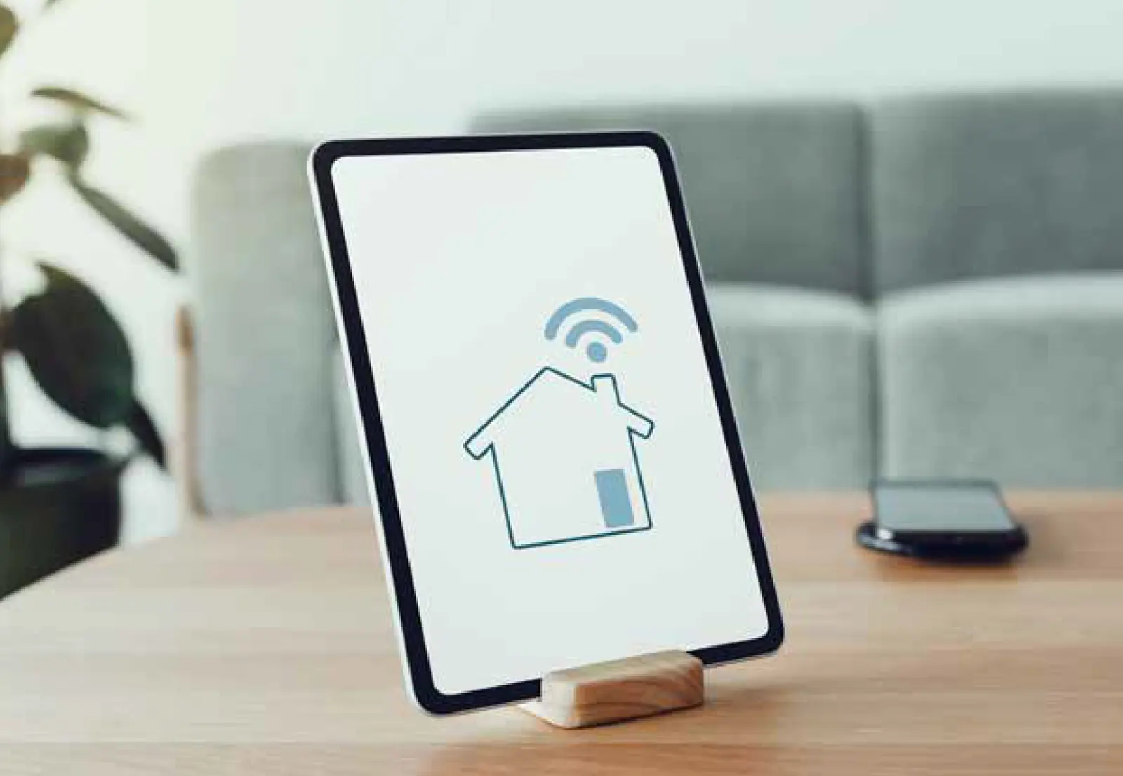 Ein Tablet in einer Halterung auf einem Holztisch zeigt ein Haus-Symbol mit WLAN-Signal. Im Hintergrund ist ein modernes Wohnzimmer angedeutet. Visualisierung von intelligenter Gebäudetechnik.